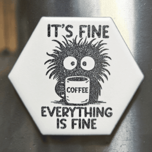 A white ceramic hexagon tile magnet engraved with a frazzled creature holding a coffee mug and the words It's Fine Everything Is Fine