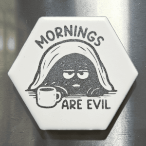 A white ceramic hexagon tile magnet engraved with a grumpy creature under a blanket holding a coffee mug and the words Mornings Are Evil