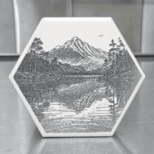 A white hexagon mountain lake ceramic magnet featuring a detailed black illustration of a mountain peak reflected in a forest lake