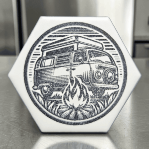 A white hexagonal camper van tile magnet with a dark circular laser-engraved image of a vintage camper van parked near a campfire.