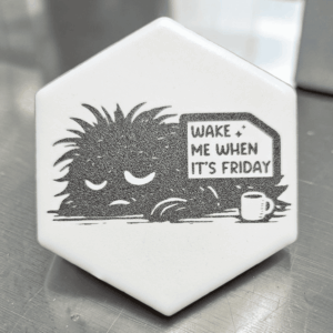 A white hexagonal funny tile magnet featuring an engraved, sleepy creature saying Wake Me When It's Friday next to a coffee mug.